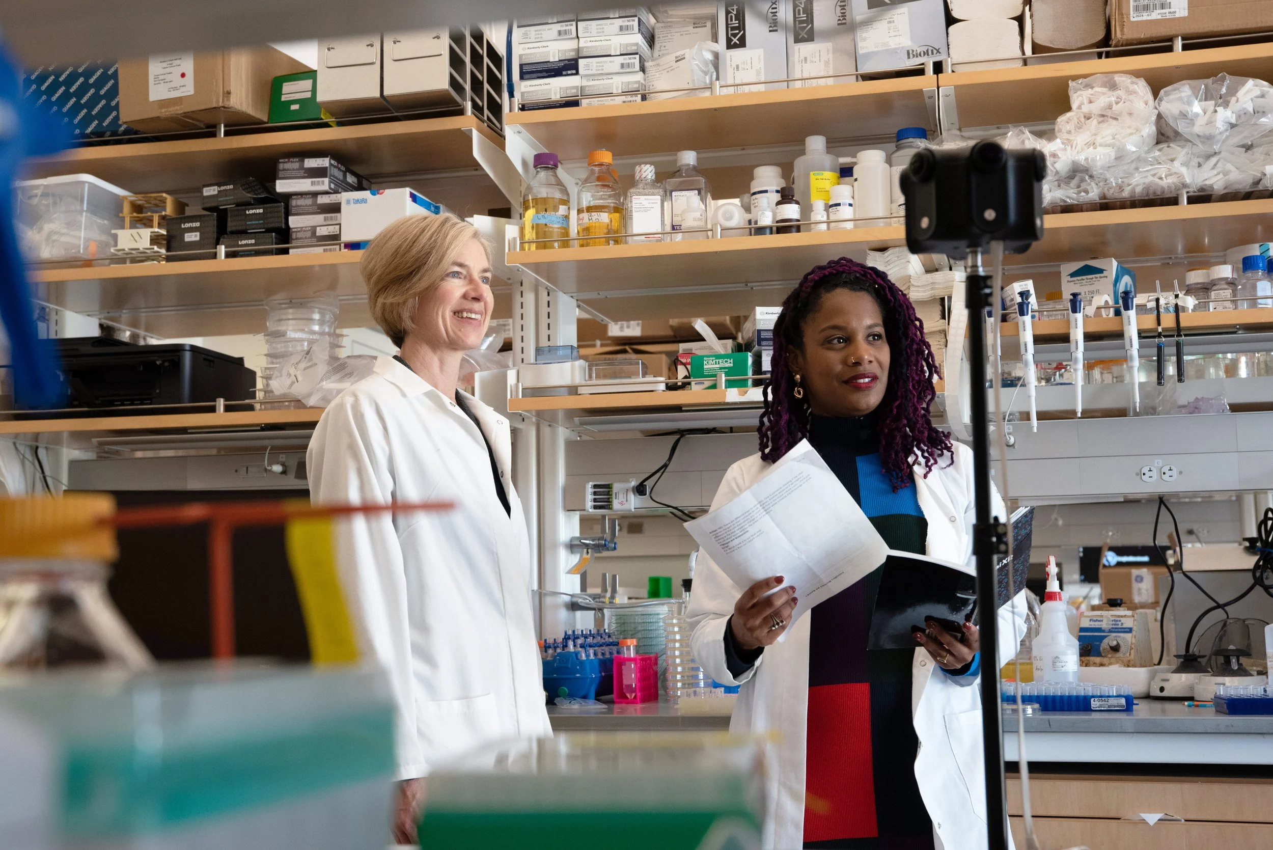 Two female scientists in a laboratory setting, one with short blonde hair smiling and the other with purple dreadlocks holding papers, seated in front of a camera on a tripod.
