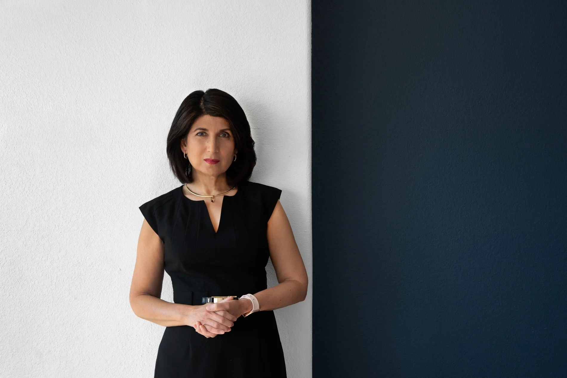 A woman with dark hair wearing a black sleeveless dress stands against a background with a white wall on the left and a dark wall on the right.