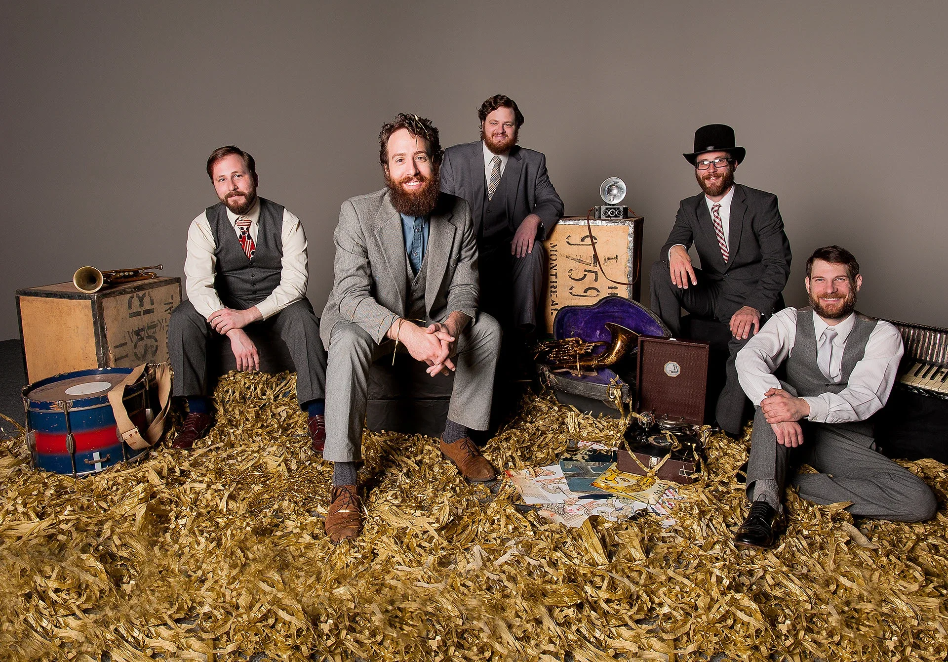 Six men in vintage suits with musical instruments and items, surrounded by gold foil, posed in a studio with a plain background.
