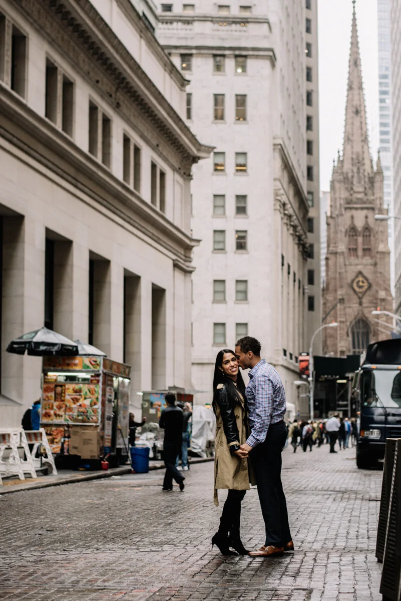 A couple holding hands on a city street, smiling, with tall buildings and a church steeple in the background.