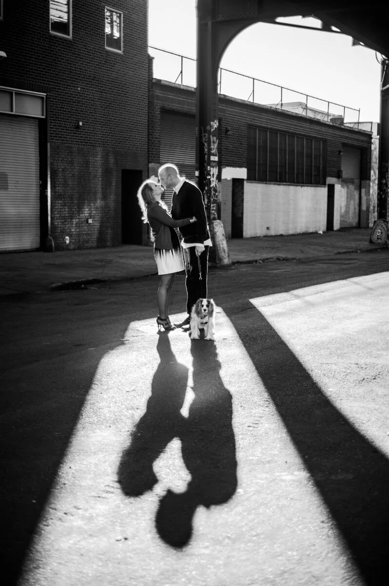 A man and woman standing close together with a dog on a city street under an overpass, captured in black and white with shadow play.