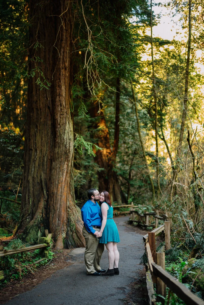 A couple standing on a trail in a lush forest during sunset, holding hands and sharing a kiss.