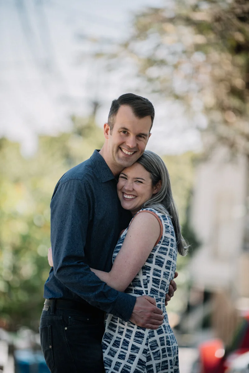 A happy couple hugging outdoors with greenery and trees in the background.