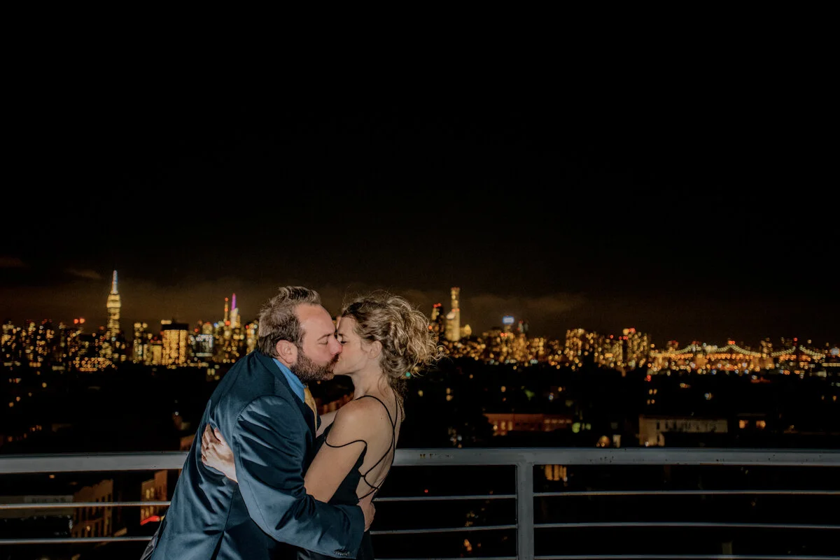 A couple kissing on a rooftop at night with a city skyline and lit buildings in the background.