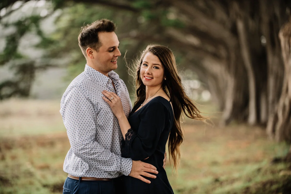A smiling woman with long brown hair and a man with short brown hair, wearing a white shirt with small black polka dots, standing close together outdoors on a tree-lined path, with the woman touching the man's chest and the man holding her waist.