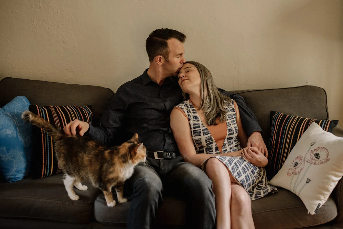 A couple sitting on a couch, with the man kissing the woman's forehead. A calico cat is on the couch near the man's lap. The woman has gray hair and is wearing a patterned dress. The couch has various pillows, including one with a floral design.
