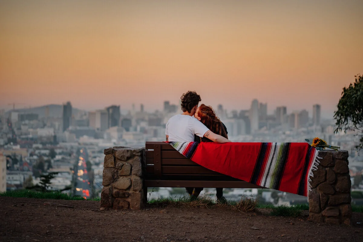 A couple sitting on a park bench overlooking a city skyline at sunset, with a colorful blanket and a sunflower beside them.