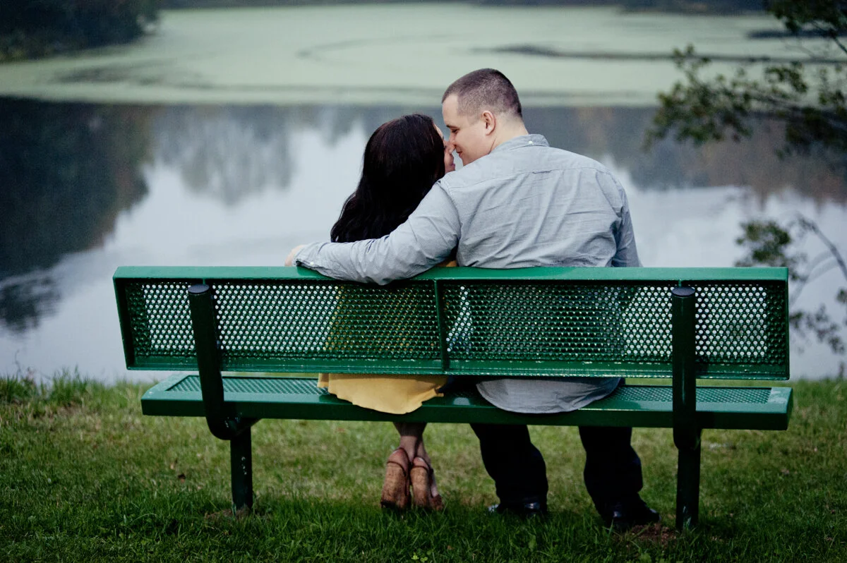 A couple sitting close together on a green park bench, leaning in towards each other, near a calm lake with trees reflecting on the water.