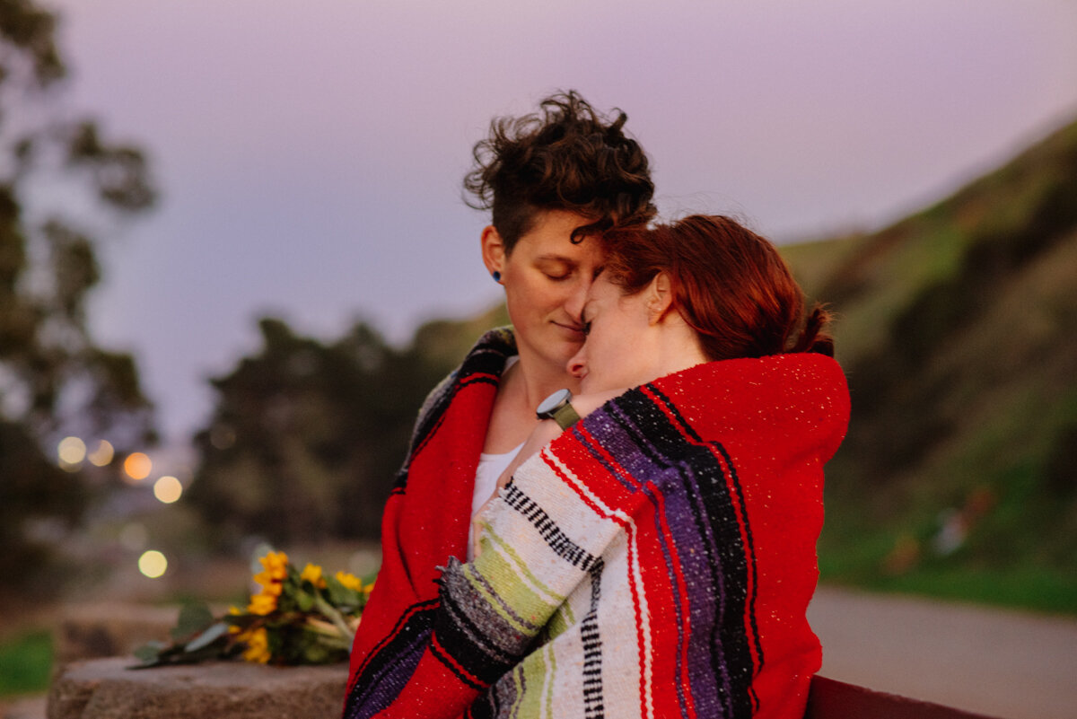 Two women embracing outdoors at sunset, wrapped in a colorful blanket, with a blurred background of trees and hills.