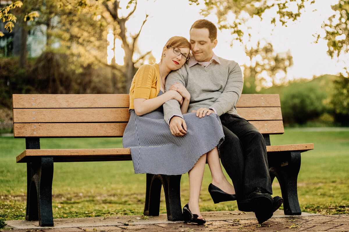 A couple sitting closely together on a park bench, embracing each other with content expressions during sunset.