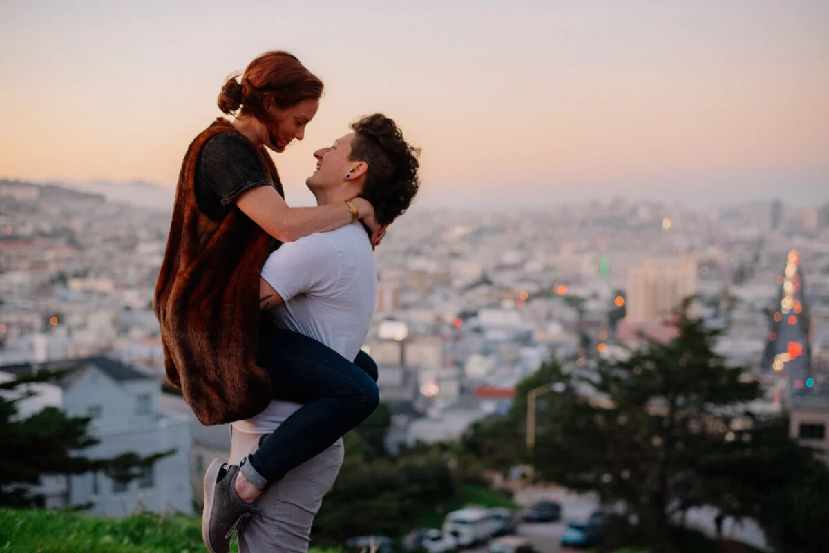 A couple sharing a joyful moment outdoors, with one lifting the other, cityscape at sunset in the background.