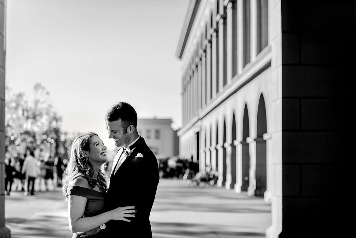 A couple in formal attire sharing a joyful moment outdoors near a large building, with other people in the background, captured in black and white.