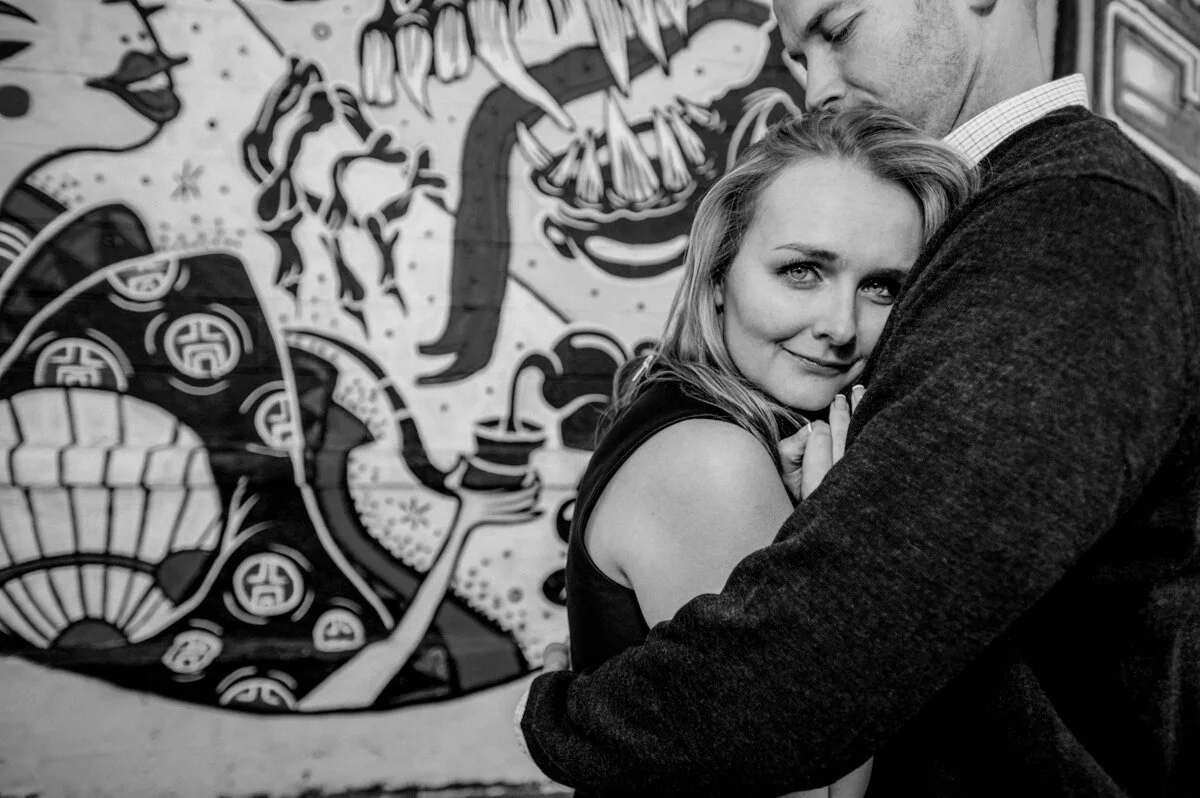 A black-and-white photo of a woman and man embracing in front of a colorful mural.