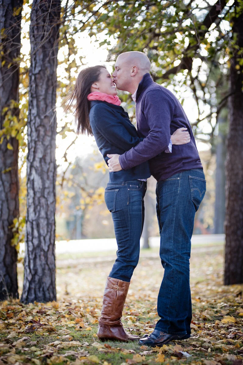A couple kissing outdoors in a park on fall day, surrounded by trees with autumn leaves.