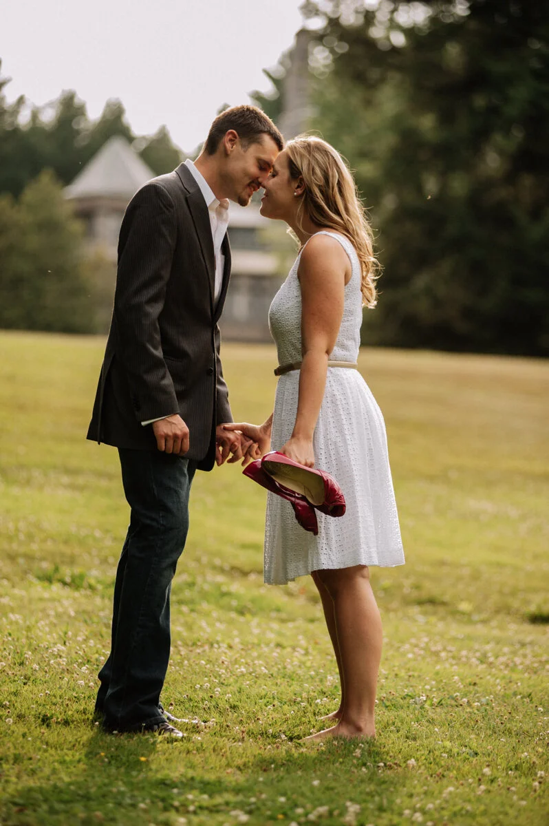 A couple standing outdoors on a grassy field, touching foreheads and holding hands, with the woman holding a red handbag, and trees and a building in the background.