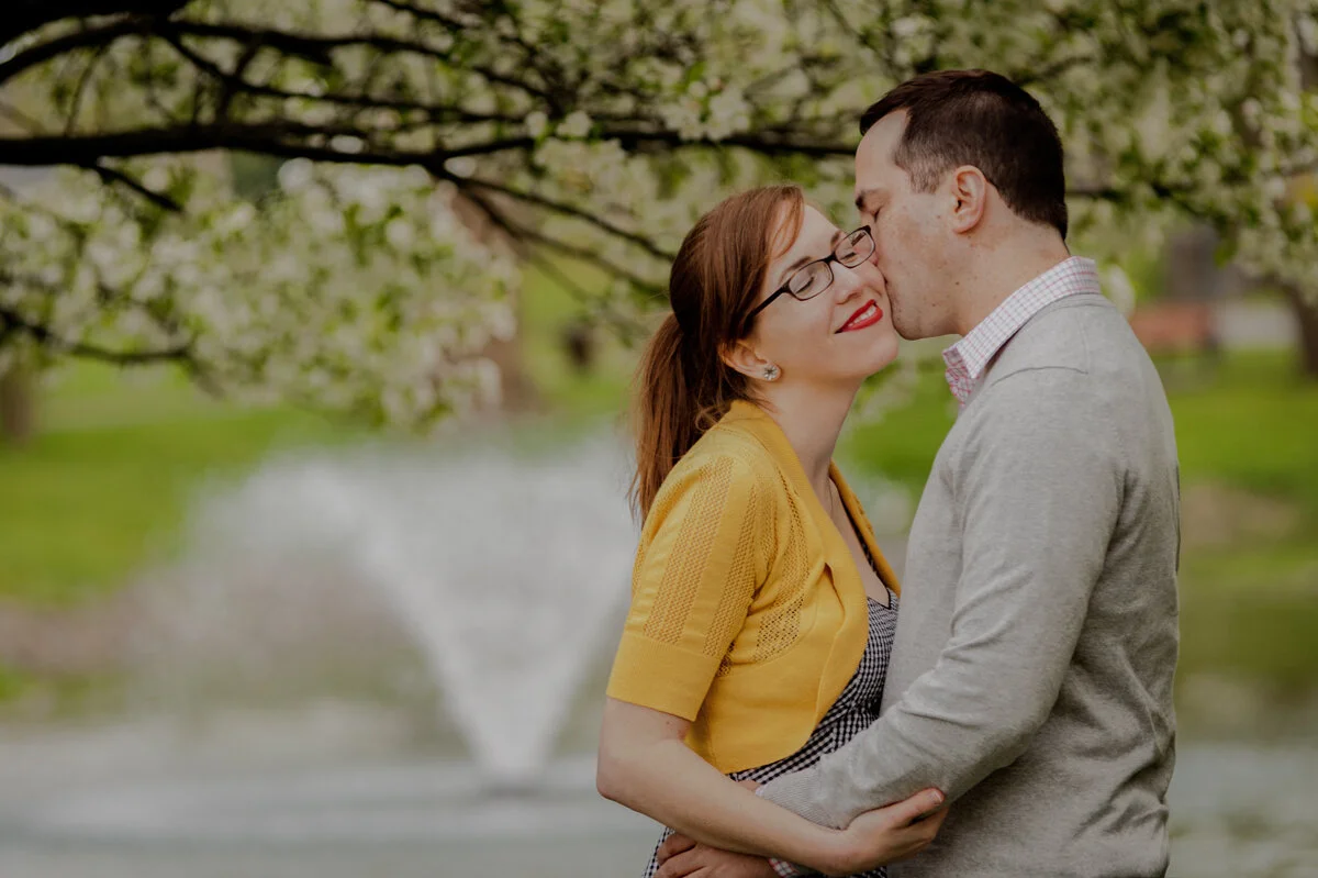 A couple stands close together by a pond with a fountain, surrounded by trees with white blossoms, in a park on a spring day.