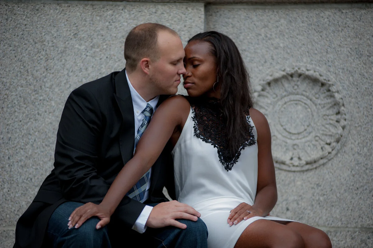 A young man and woman sitting closely face to face against a stone wall, about to kiss.