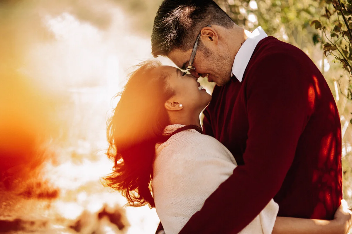 A couple is embracing outdoors with their foreheads touching, smiling, and looking into each other's eyes during sunset.