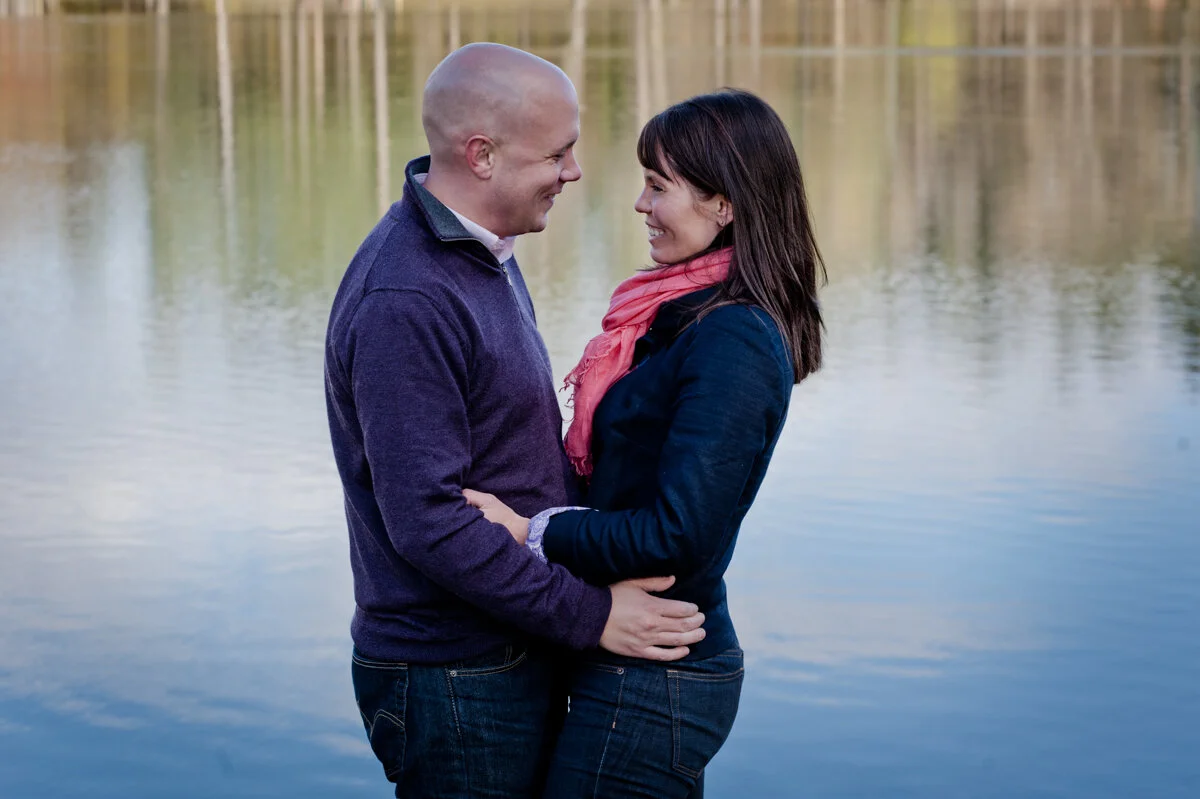 A couple standing close together by a lake, smiling and looking into each other's eyes during daytime.