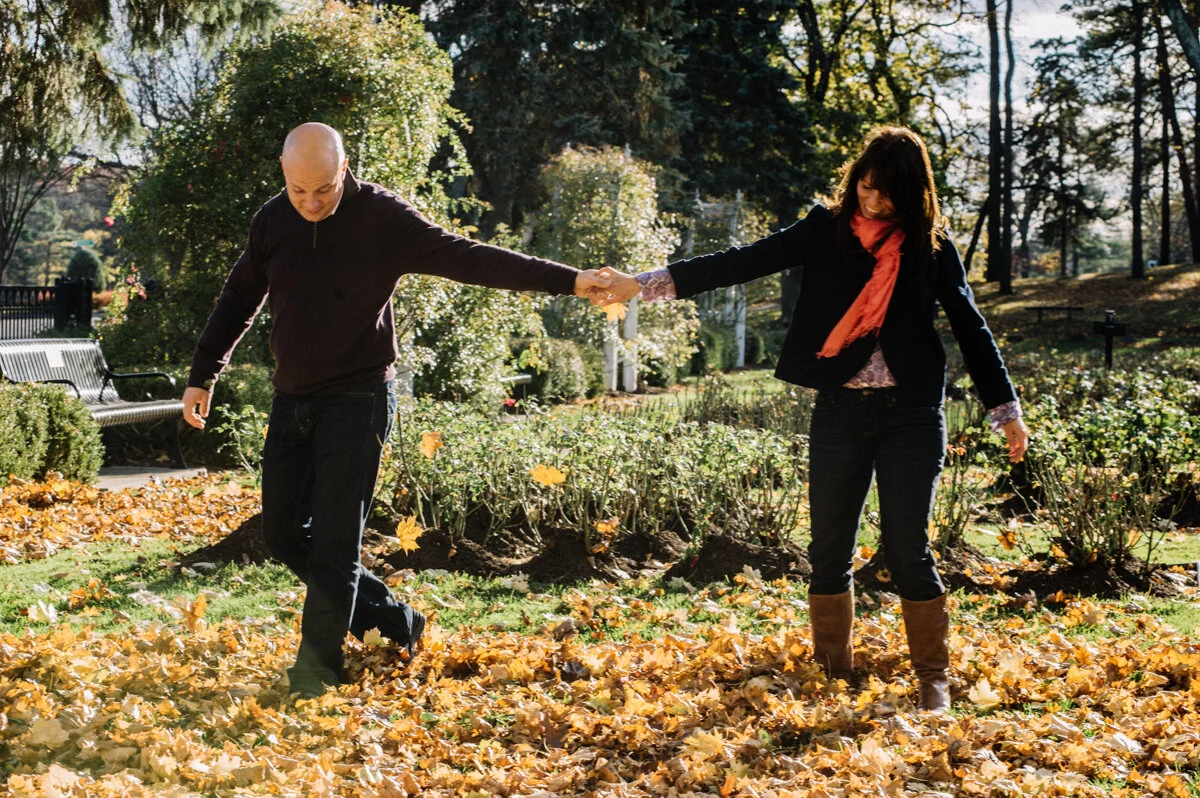 A man and woman holding hands in a park during fall, surrounded by fallen leaves and trees.