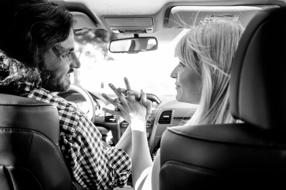 A black-and-white photo of a man and woman sitting inside a car, facing each other, smiling, and holding hands.