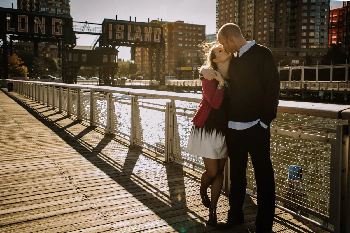 A couple kissing on a wooden dock by a river with buildings and a bridge in the background, during sunset.