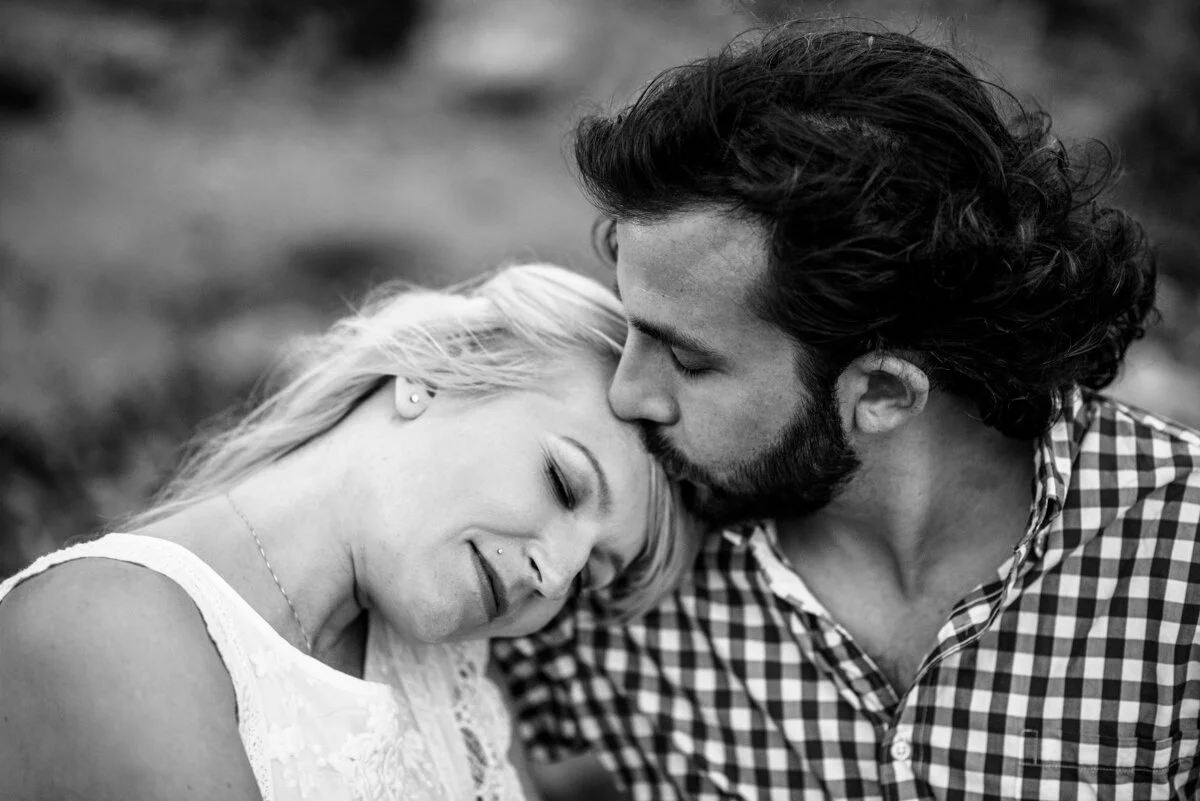 A black-and-white photo of a man kissing a woman on the forehead, both with eyes closed and smiling, outdoors.