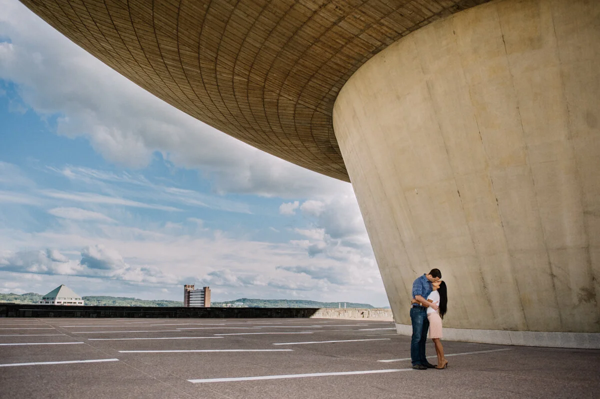 A couple sharing a kiss under a large, modern, concrete and wooden architectural structure in an empty parking lot with a cityscape and cloudy sky in the background.