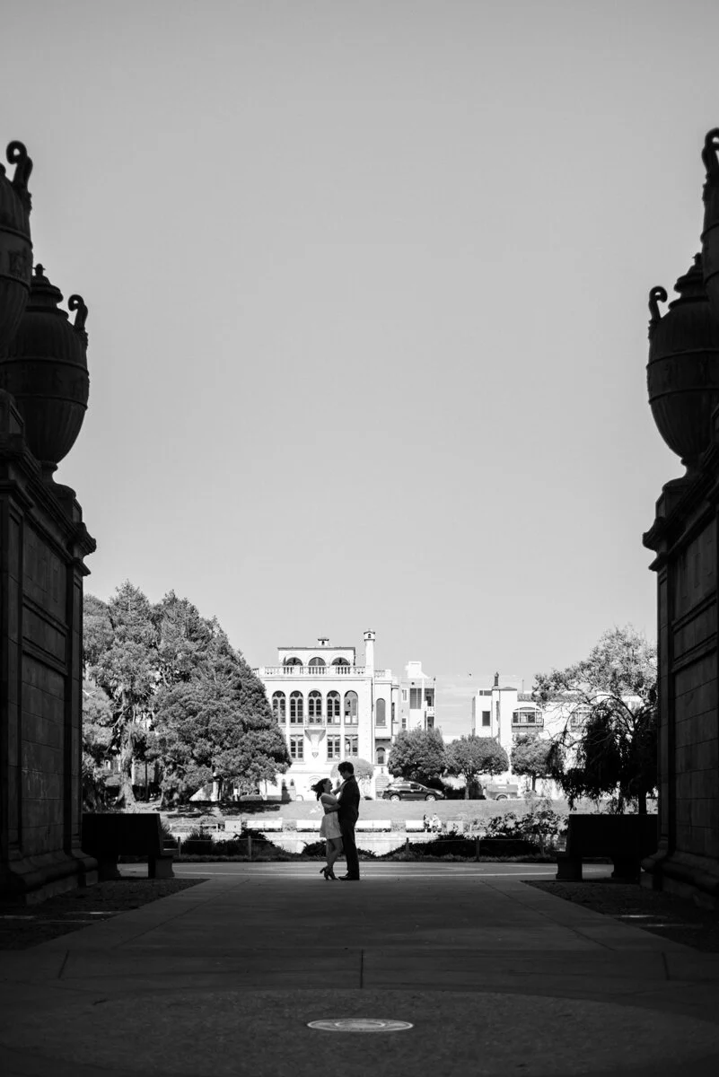 Silhouetted couple dancing in a park, seen through an archway with trees and buildings in the background, black and white photograph.