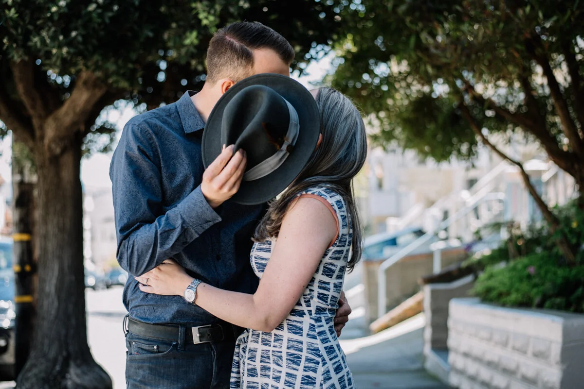 A man and woman sharing a kiss outdoors, with the man holding a black hat over their faces, standing under a large tree on a sunny day.