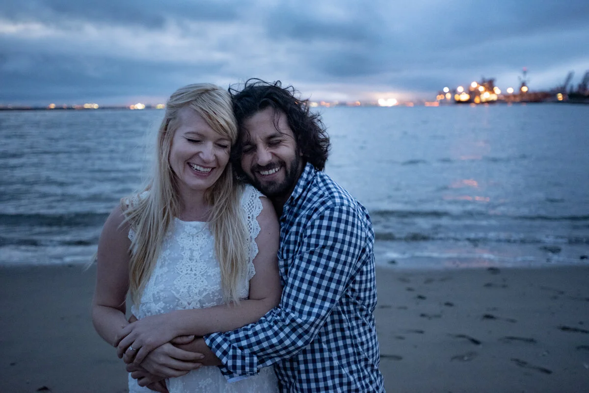 A smiling couple hugging on a beach at dusk with city lights in the background.