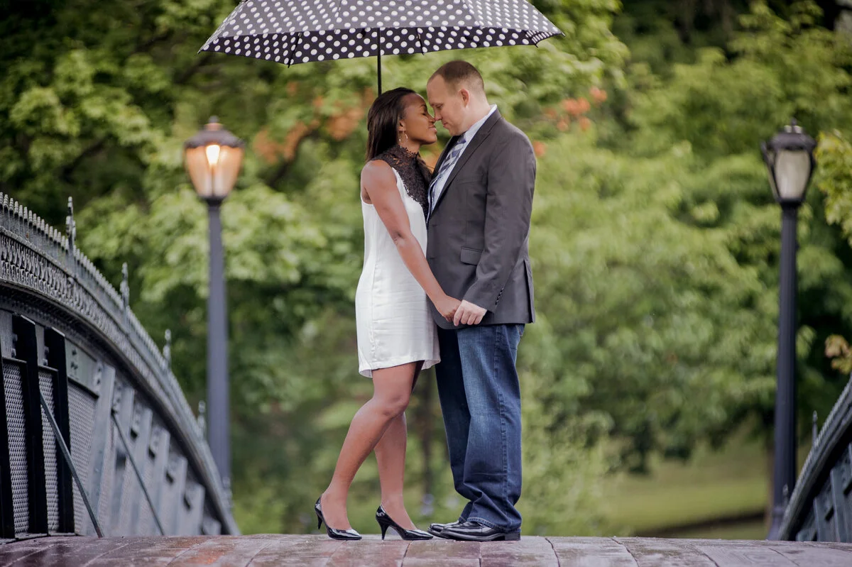 A couple standing on a bridge under a black umbrella with white polka dots, holding hands, with green trees in the background.