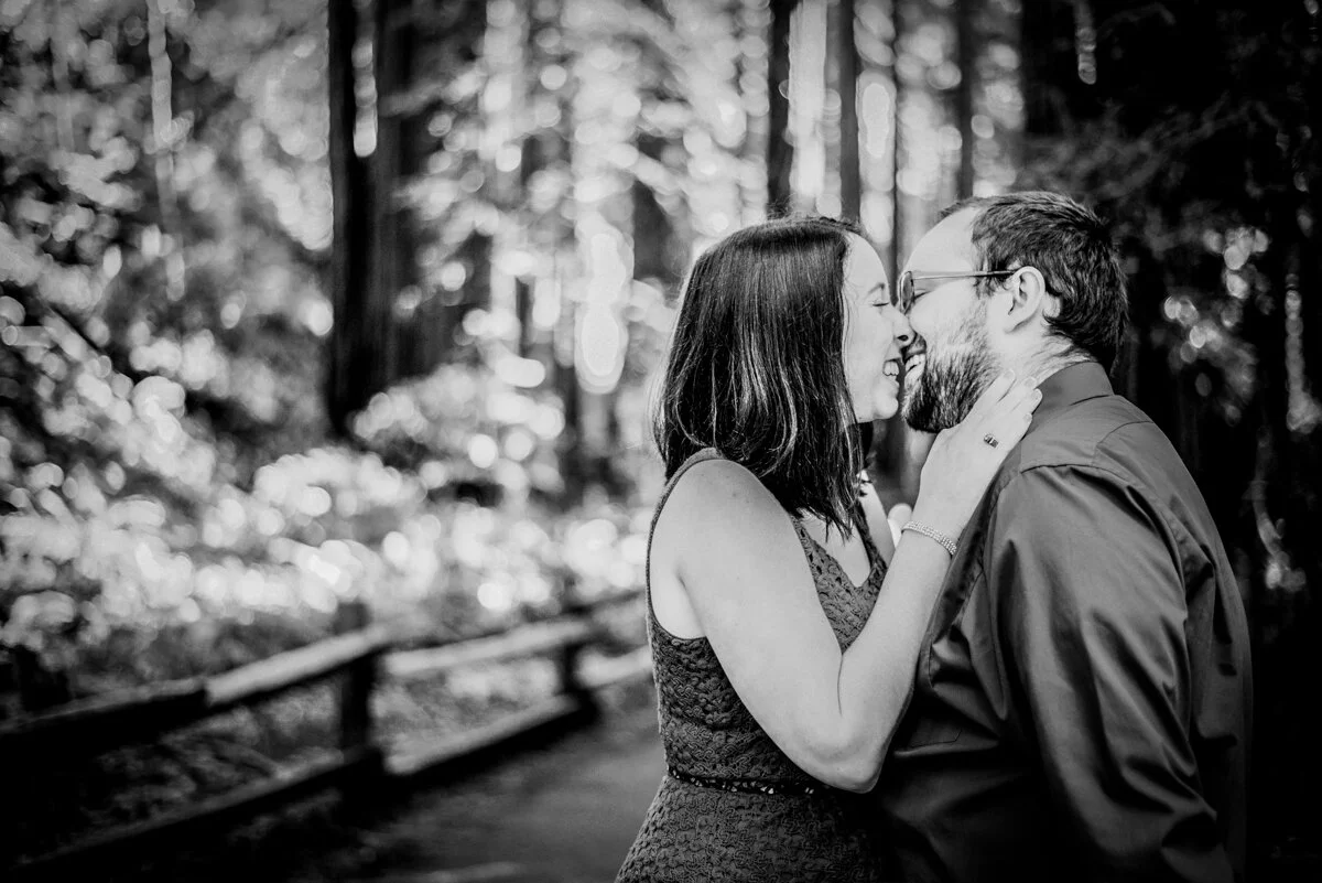 A couple sharing a kiss and smiling in a forest setting with trees in the background.