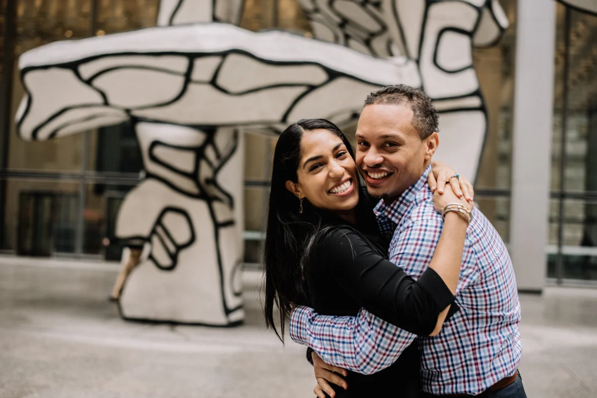 A smiling couple hugging in front of a large black and white mushroom sculpture outdoors.