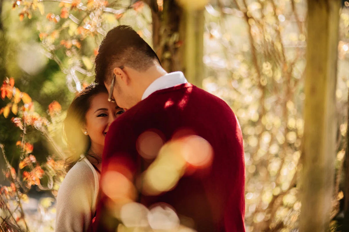 A couple in a forest, with the woman smiling as she looks at the man, who is facing away from the camera. The scene is illuminated by warm, golden light.
