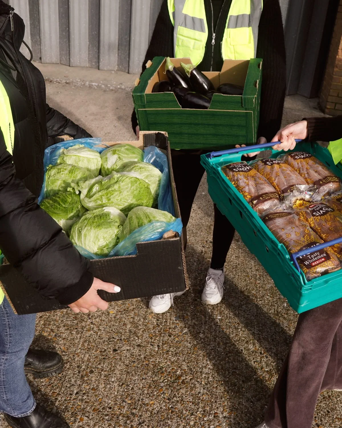 Volunteers holding boxes of leafy lettuce, eggplants, and packaged baked goods outside.