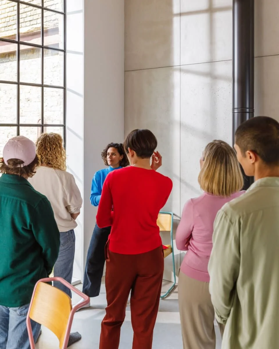 Group of people listening to a woman speaking during a presentation or discussion in a bright, modern room with large window and concrete walls.