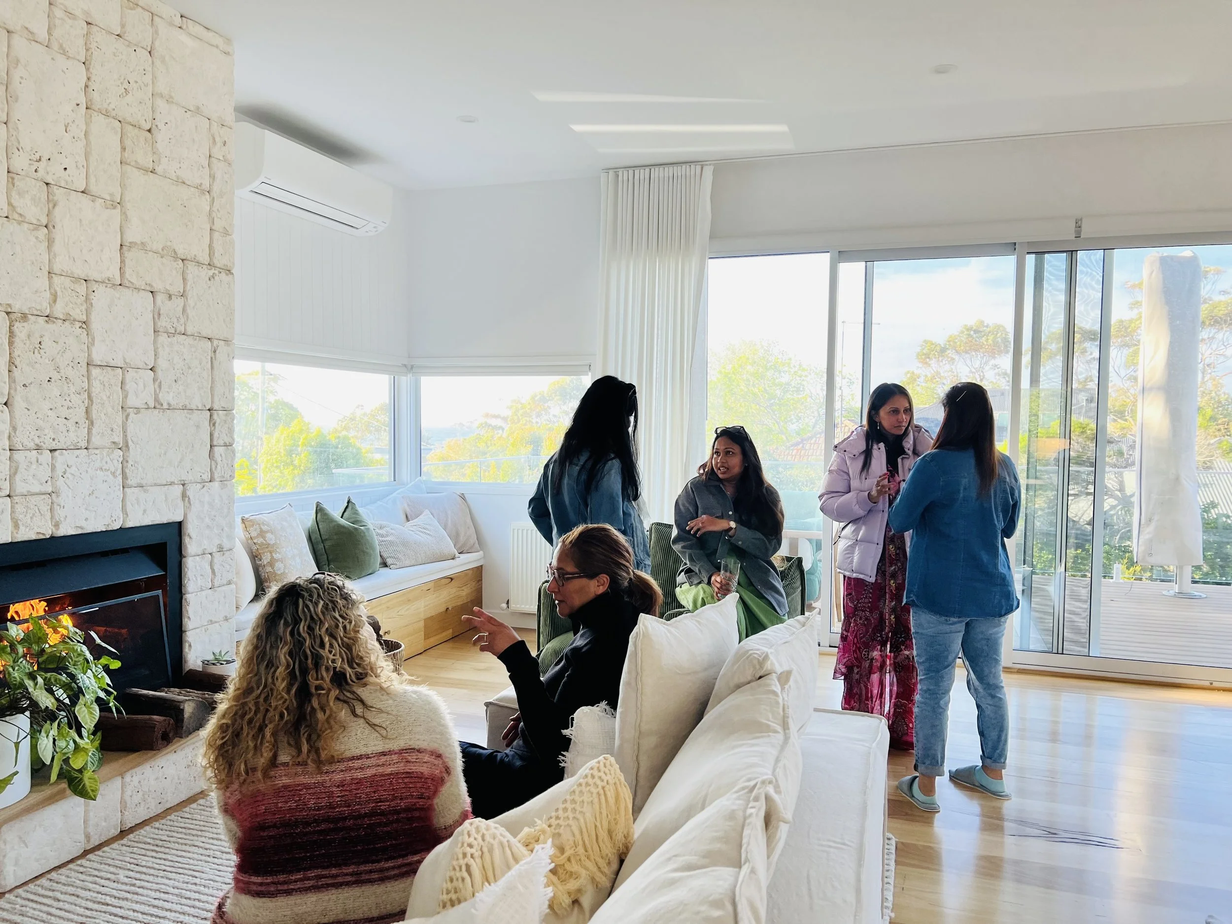 Group of women talking and socializing in a bright living room with large windows and wooden flooring.