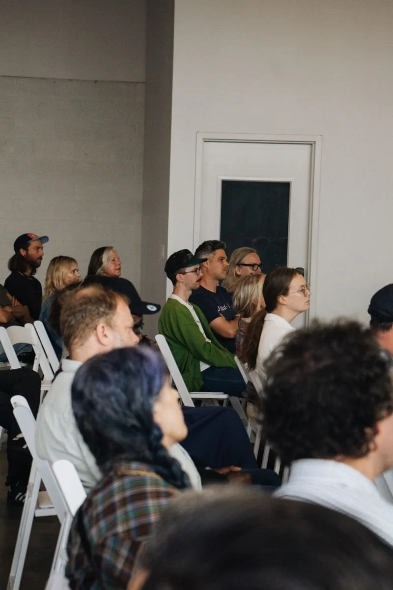 An audience seated on white chairs in a room, attentively listening to a speaker or presentation, with mostly young adults and a diverse group of people.