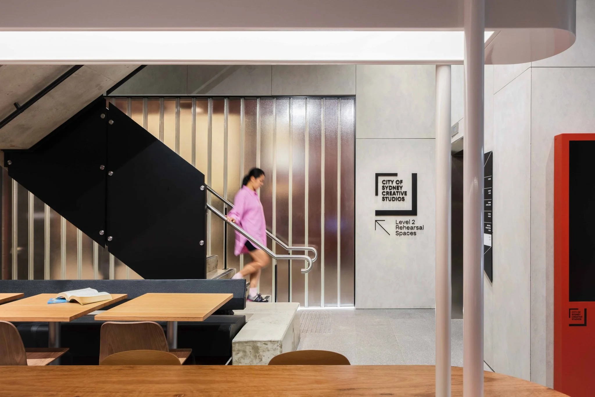 Interior of a modern building with wooden tables and chairs, a staircase with a woman in a pink coat walking up, and signs indicating a creative studios space and rehearsal areas.