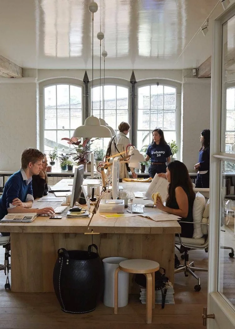 People working and talking in a bright, modern office with large windows, desks, and plants.