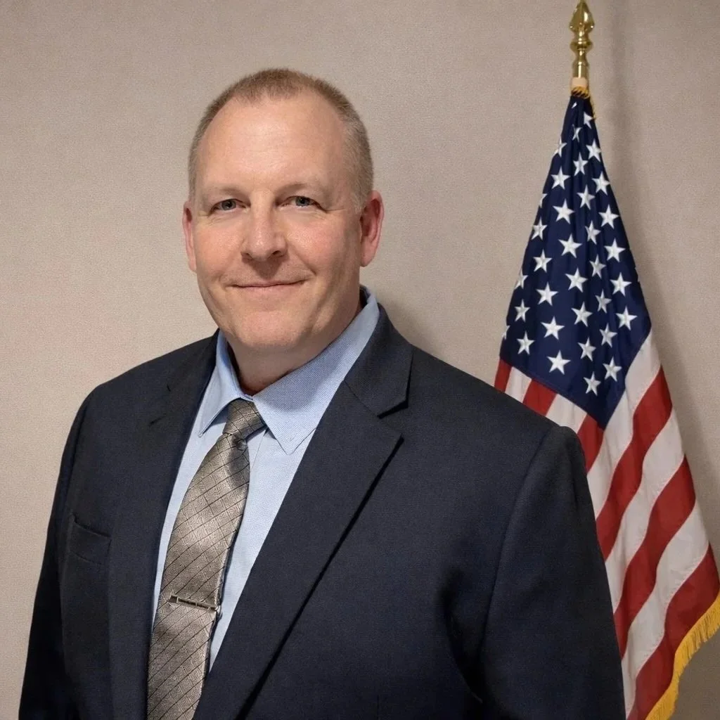 A man in a dark suit and tie standing in front of an American flag.