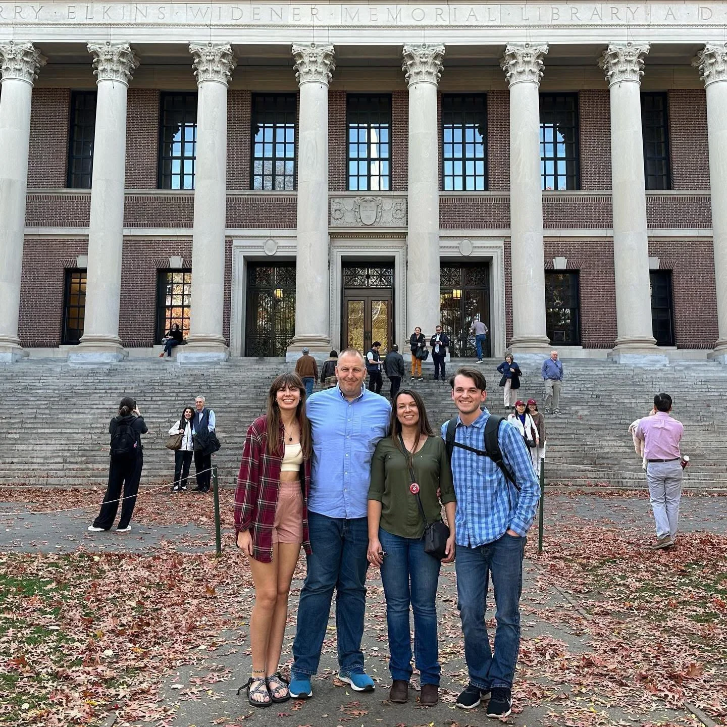 Don Burrows with his family standing in front of Widener Memorial Library, part of Harvard University, with steps and brick facade visible. Others are seen on the steps or walking. Fall leaves cover the ground.