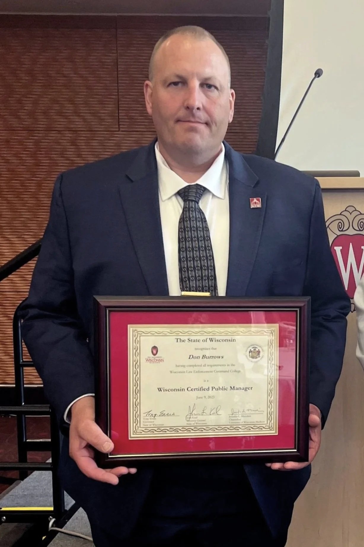 A man in a dark suit and patterned tie holding a framed certificate of recognition from the State of Wisconsin for a Wisconsin Certified Public Manager.