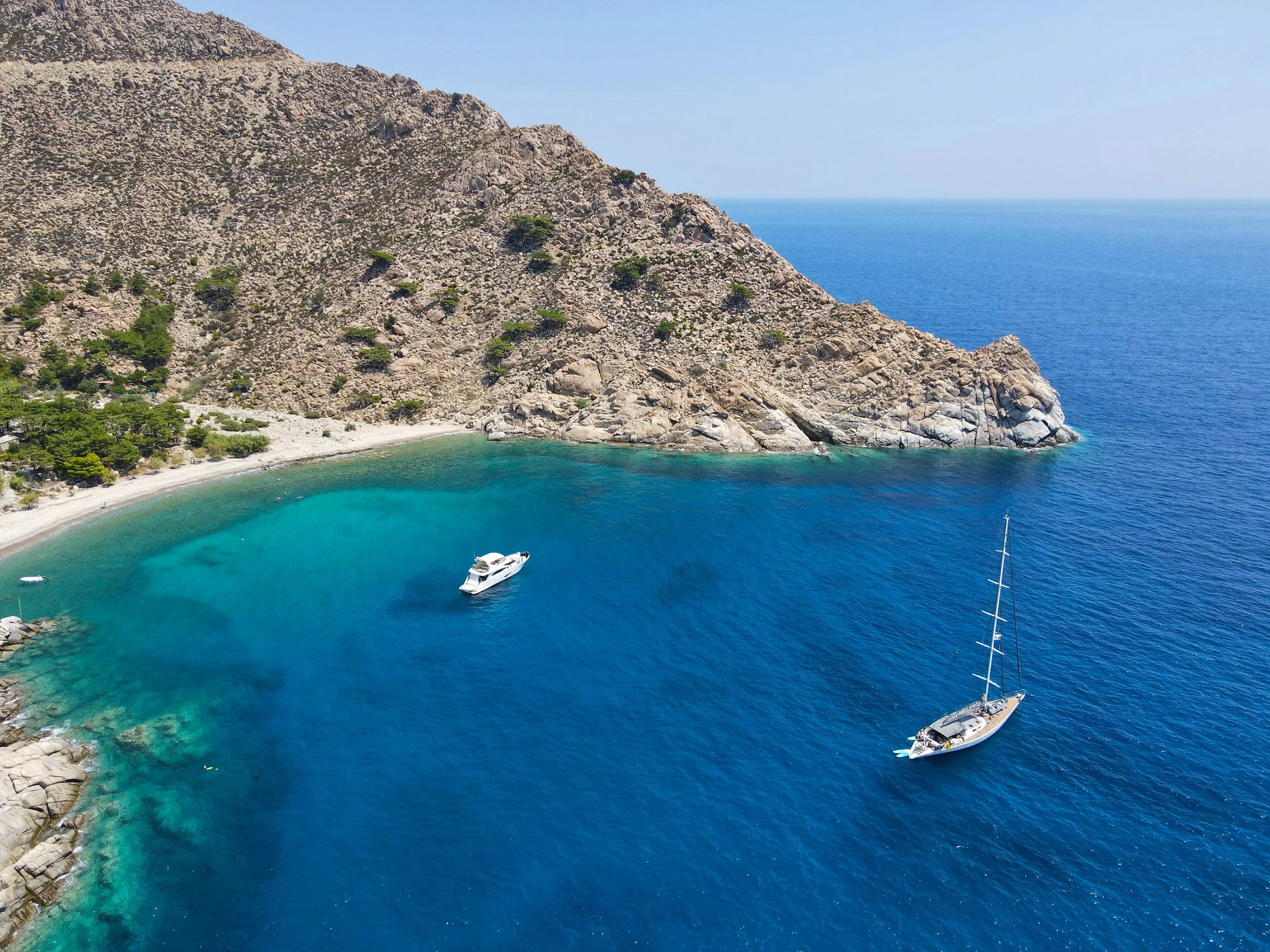 Sailboats anchored in a clear blue Aegean bay beside a rocky Greek coastline.