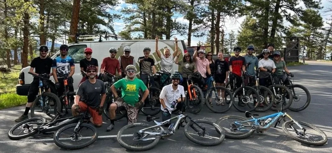 Group of people with bicycles outdoors near a van in a forested area, smiling and posing for a photo.