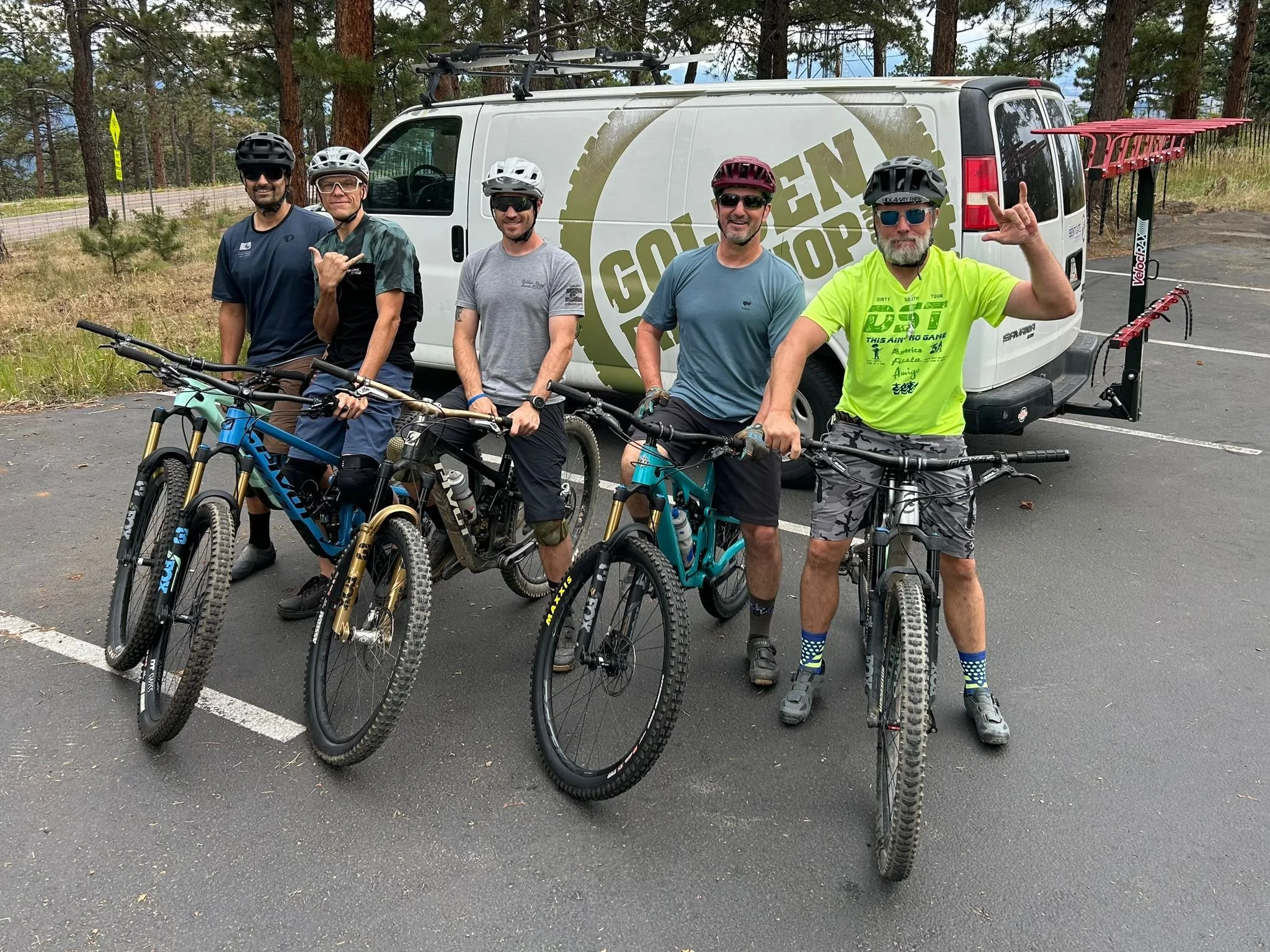 Six men with mountain bikes and helmets standing in a parking lot, with a white van featuring a logo behind them, surrounded by trees.