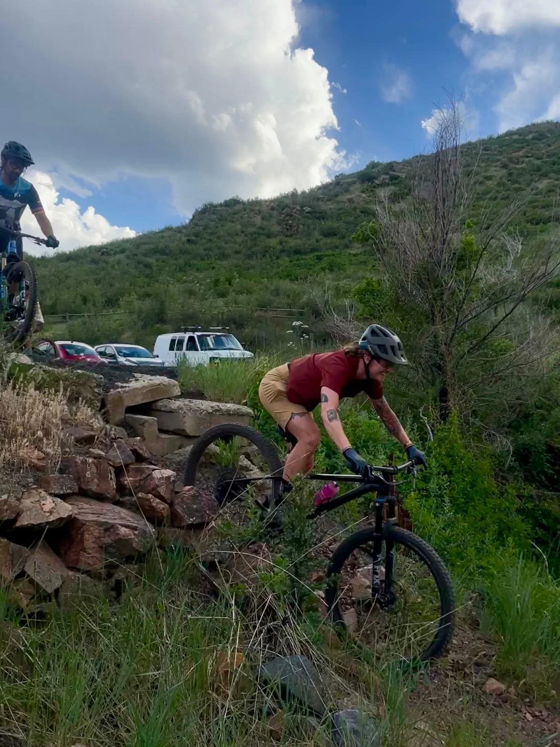 A person riding a mountain bike down a rocky trail in a green outdoor area with hills and cloudy sky in the background.