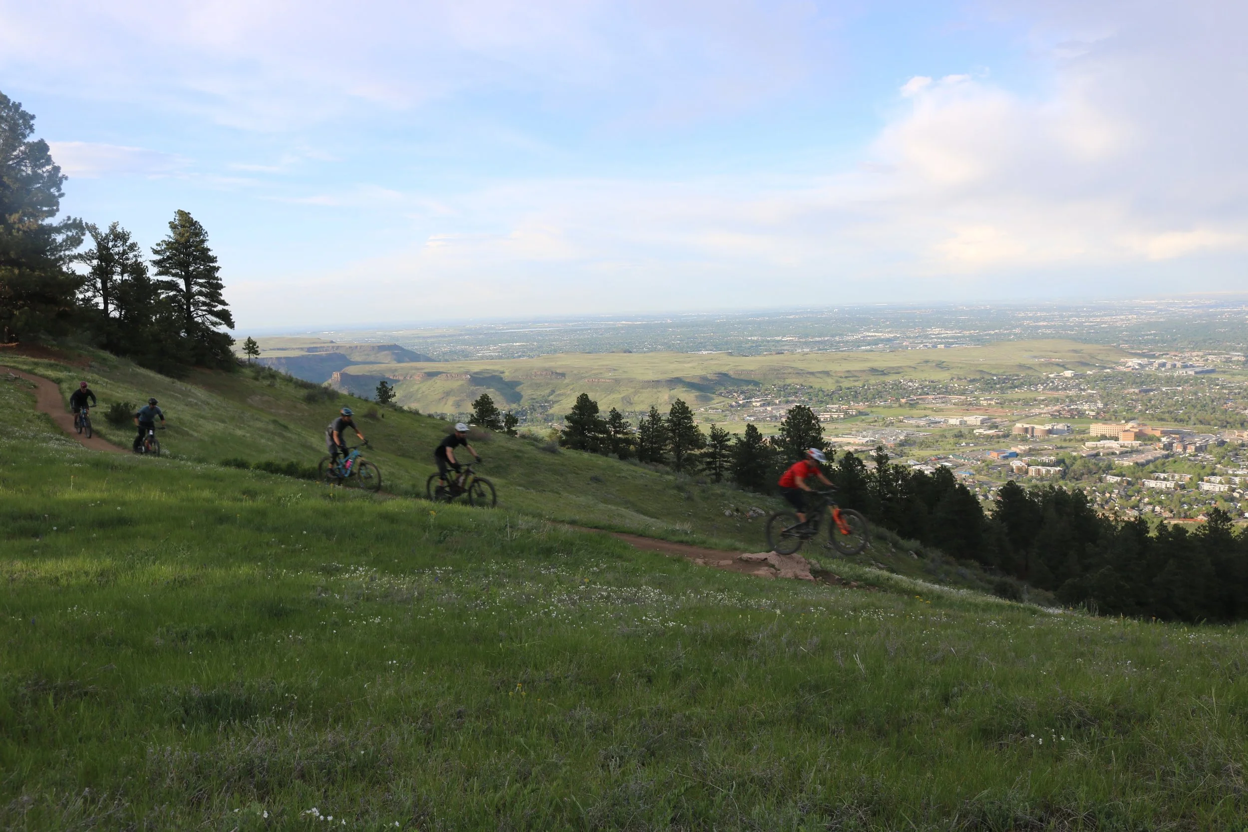 Six mountain bikers riding on a trail down a grassy hill with trees, overlooking a cityscape and distant mountains under a partly cloudy sky.