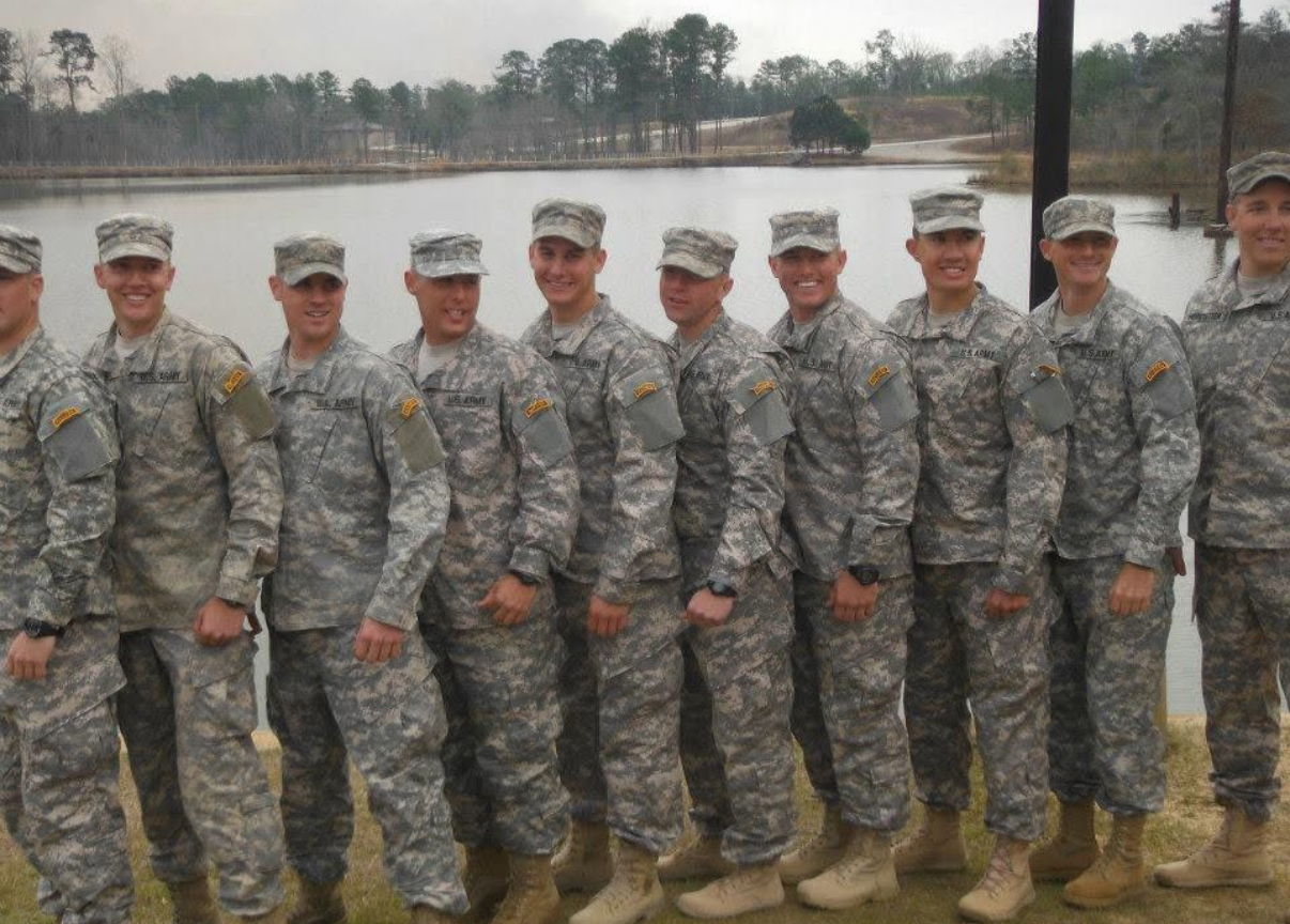 A group of U.S. Army soldiers in uniform standing outdoors near a body of water, smiling for a group photo.